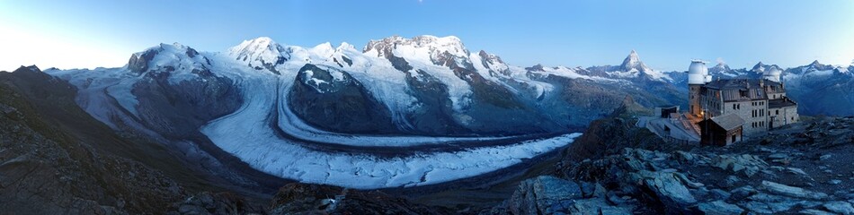 A panorama of Gorner Glacier with the moon shining over snow capped mountains (Monte Rosa Massif, Liskamm & Matterhorn) and the Gornergrat Observatory on a rocky ridge in Zermatt, Valais, Switzerland