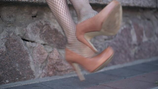 Slim female legs in fishnet stockings taking off brown high-heels outdoors. Unrecognizable young Caucasian woman in uncomfortable shoes in urban city. Lifestyle and femininity concept