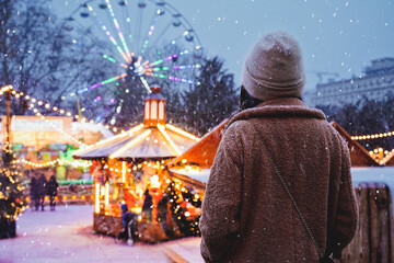 Woman looks at night Christmas city. Girl on background of carousels and amusement park in winter