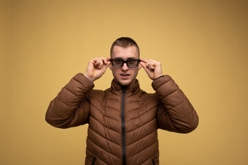 Obraz premium A young man of the 20s in a brown jacket, on a yellow background, adjusts his glasses to watch a 3d movie