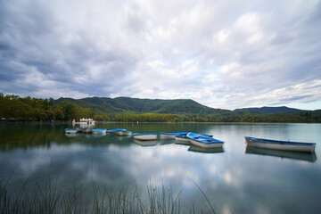 Boats stranded in the calm waters of Banyoles Lake with a bathhouse and the mountains in the background on a cloudy sunrise. Largest natural lake in Catalonia, Spain, Europe.