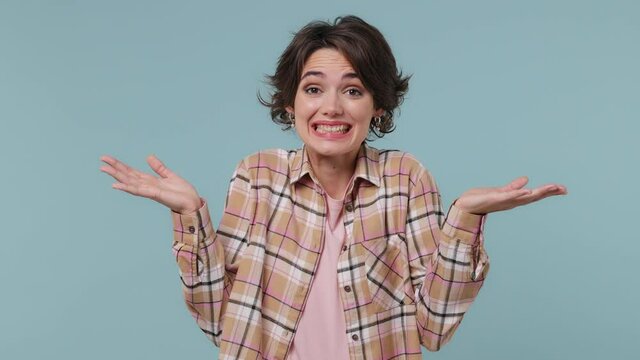 Fun confused shy shamed young brunette woman 20s wears plaid shirt looking camera spreading hands say oops ouch oh omg i am so sorry isolated on pastel plain light blue background studio portrait