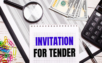 On the office table there is a notebook with the text INVITATION FOR TENDER, a pen, a calculator, dollars, multi-colored paper clips and a magnifying glass. Stylish workplace. Business concept