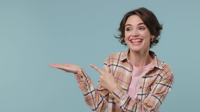 Happy promoter vivid young brunette woman 20s wears plaid shirt pointing fingers aside away on empty palm with copy space place mock up isolated on pastel plain light blue background studio portrait