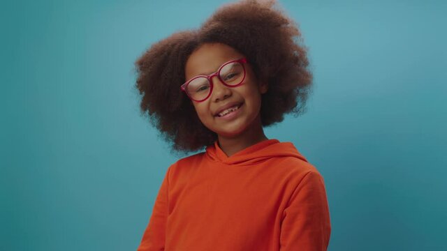 Smiling African American girl in eye glasses wearing orange clothes looking at camera on blue background. 7 years old happy kid in glasses.