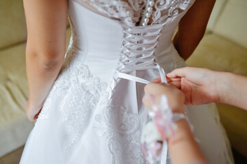 Obraz na płótnie Canvas The bridesmaid prepares the bride for the wedding day, helps to button and tie her dress. Bridesmaid makes a bow on the back of the bride's wedding dress, the bride's morning gathering