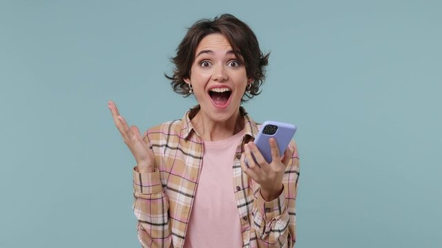 Surprised joyful young brunette woman 20s wears plaid shirt hold use mobile cell phone typing say wow just found out great big win news isolated on pastel plain light blue background studio portrait