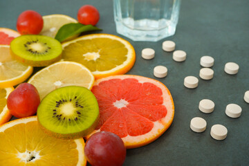 Supplements or healthy natural food? Scattered white pills vitamins, Sliced citrus fruits, orange, lemon, kiwi, grapes, glass with water on a stone gray background