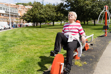 Senior woman with eyeglasses doing exercise on static bicycle machine at park on sunny day. Elder...