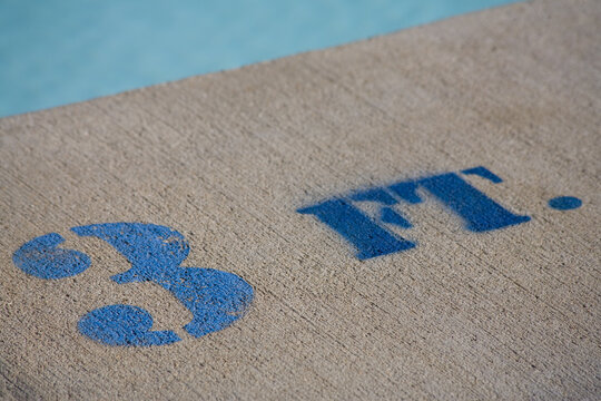 The painted sign on the concrete deck of a swimming pool spells 3 FT. This close-up shows water in the background.