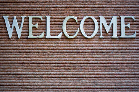Display case sign that spells out WELCOME. Close-up, full frame, white and faded white letters on a pinkish gray background.