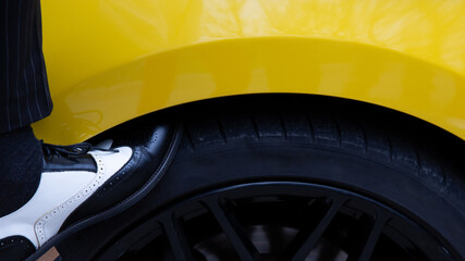 A man in beautiful classic shoes rests his foot on the wheel of a yellow car - in the frame the leg and part of the wheel