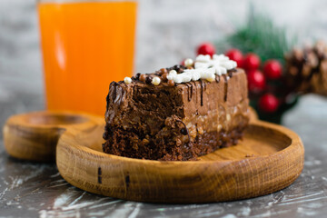 Delicious mousse chocolate brownie in a cut on a wooden plate, a glass of juice and Christmas decor on the background.