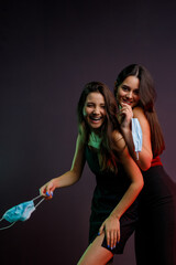 Naklejka premium Two exited young women in evening dresses and manicure happy having fun and keeping in their hands medical masks to go to the party on pandemic time.
