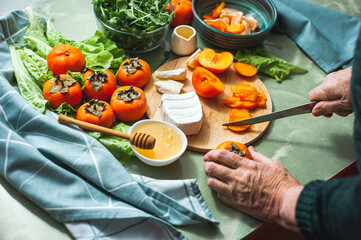Elderly vegan woman prepares a sweet salad of persimmons, tangerines and soft cheese with honey....