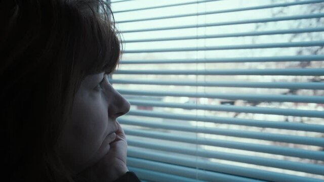 close up of pensive middle-aged woman looking out the window with blinds, looking around, waiting for someone