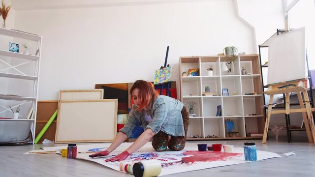 Creative inspiration. Happy female painter. Art studio. Cheerful woman using hands for painting white map paper sitting floor light room interior.