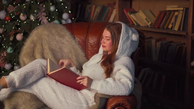 Christmas. Woman reading book sitting cozy leather armchair with Christmas tree in background . Beautiful young caucasian female resting home reads book dressed warm pajamas jumpsuit