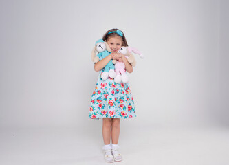 A little beautiful girl in a bright dress hugs two favorite toys on a white isolated background.