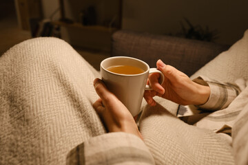 A cup of tea in the hands of a girl. The girl is drinking hot tea. A girl in pajamas is wrapped in a blanket and is drinking delicious tea. Enjoy the comforts of home. White cup close up in woman hand