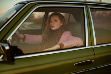 Fashionable confident woman posing at the wheel of green retro car. Elegant redhead model wearing...