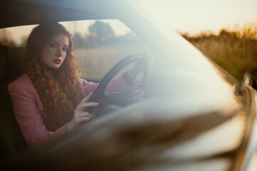 Fashionable confident woman posing at the wheel of green retro car. Elegant redhead model wearing...