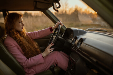 Fashionable woman posing at the wheel of green retro car. Elegant redhead model wearing classic pink suit. Copy, empty space for text