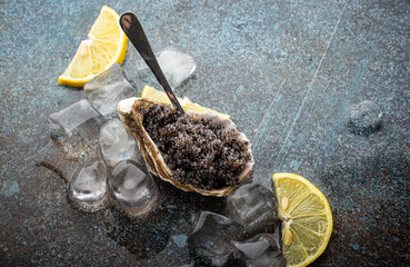 Black caviar served in oyster shell with spoon with lemon wedges and ice cubes on rustic stone blue...