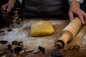 A rectangle of dough lies on baking parchment, a rolling pin and cookie cutters are lying next to it, everything is strewn with flour, a cook is standing in the background and holding a rolling pin