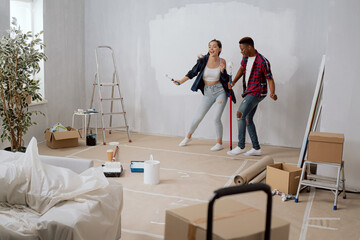 Entertaining cheerful friends spend time together, have fun while renovating the room, apartment, hold paint rollers in their hands, paint walls with white paint dancing singing joy of buying flat
