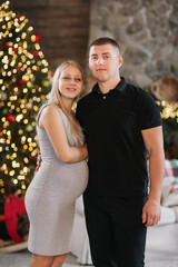 Fototapeta premium Adorable marriage couple near Christmas tree and fireplace at home, caring husband hug beautiful pregnant wife, young family enjoy happiness, winter holidays concept