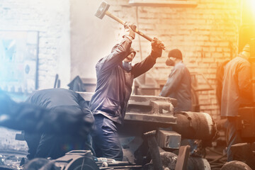 Worker swings large hammer in workshop of metallurgical plant.