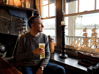 Middle aged man is sitting by the window and drinking beer in old pub and looking at rain through...