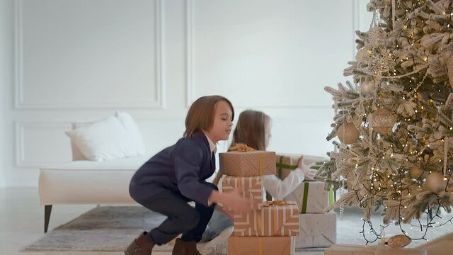 New Years Eve. Two Cute Children Put Boxes Of Gifts Under Christmas Tree. Girl With Long Hair And Cute Boy. Bright Christmas Tree.