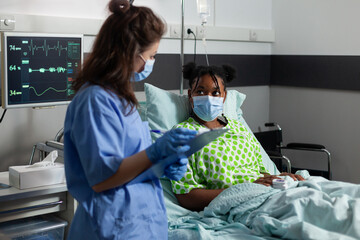 African american young patient with protection face mask against covid19 lying in bed explaining...