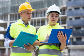 Male and female engineers standing in construction area and discussing. Woman using laptop and man holding folder in hands.