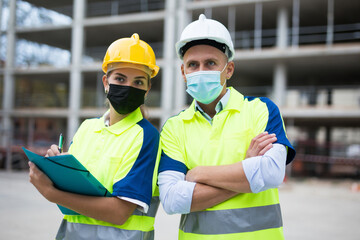 Man and woman engineers in face masks standing in construction area and looking in camera.