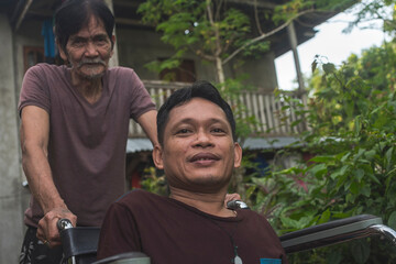 A disabled middle aged Filipino man on a wheelchair being assisted by an older guy, possibly his...