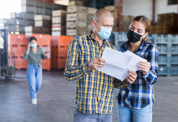Man and young woman in face masks talking about documentation while standing in warehouse. Another woman talkin on phone in background.