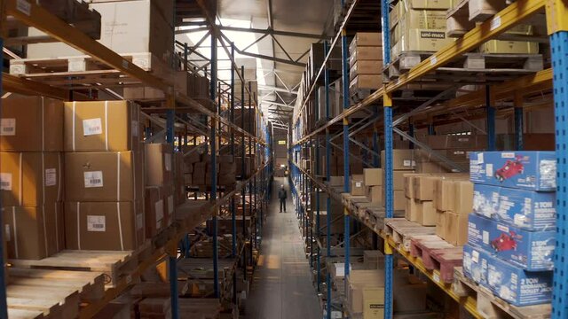 Man standing in a long storage warehouse aisle with boxes,aerial zoom.