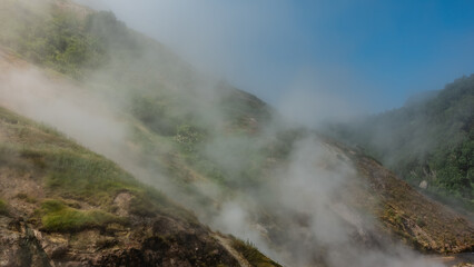 Geothermal zone of the Valley of Geysers. Kamchatka. Green vegetation grows on the slopes of the mountains. Steam from the hot springs rises into the blue sky. Close-up. Poor visibility due to haze.