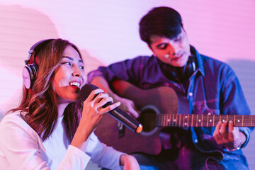Obraz premium Pretty Asian female singer recording songs by using a studio microphone with male playing guitar in blue and red light. Performance and show in the music business. Duet session.