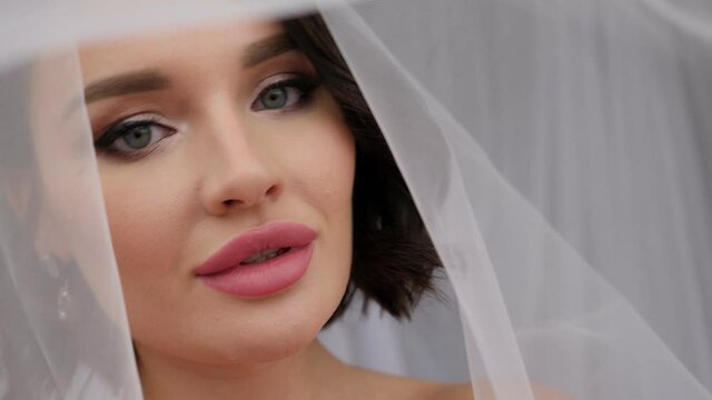 Close-up portrait of a young beautiful bride with dark hair with a head covered with a veil on a white background.