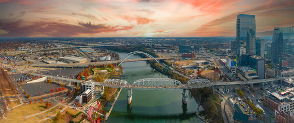 A gorgeous aerial shot of the silky green waters of the Cumberland River with bridges across the river and autumn colored trees with a vast view of the cityscape with powerful clouds at sunset - obrazy, fototapety, plakaty