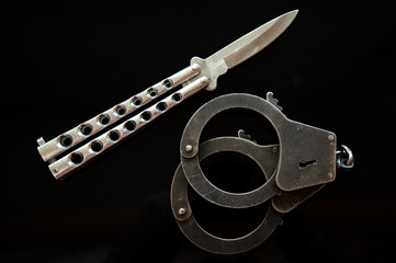 Butterfly knife on a black background. Metal handcuffs on black isolate. Criminal news. Arrest for carrying a bladed weapon.