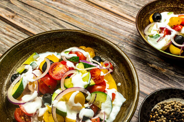 Fresh vegetable salad of fresh tomato, cucumber, onion, black olives,yellow sweet pepper with natural flavor yogurt sauce on ceramic bowl. It is located on an old natural wooden floor. Top view, copy 