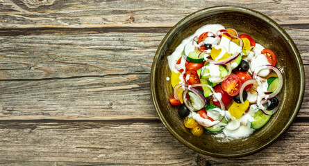 Fresh vegetable salad of fresh tomato, cucumber, onion, black olives,yellow sweet pepper with natural flavor yogurt sauce on ceramic bowl. It is located on an old natural wooden floor. Top view, copy 