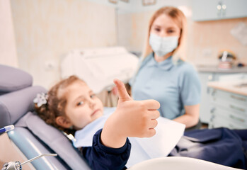 Focus on girl's hand giving thumbs up while woman stomatologist in medical mask sitting beside kid in dental chair. Concept of pediatric dentistry and dental care approval.