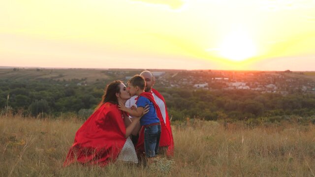 mother and father kiss and hug little child sunset, playing superheroes dressed red cloaks, happy family, brave savior costume, fantastic childhood dream heroes, fearless kid boy winner fun play game