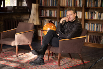 Pensive Man in a Leather Jacket and Ankle Boots Sits Cross-legged in an Old Armchair in a Vintage...